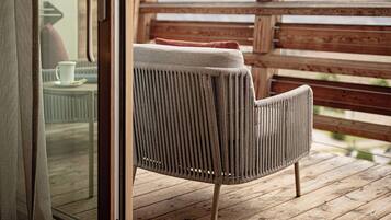 A wooden deck with a wicker chair and a glass door leading to a room with a view of mountains.