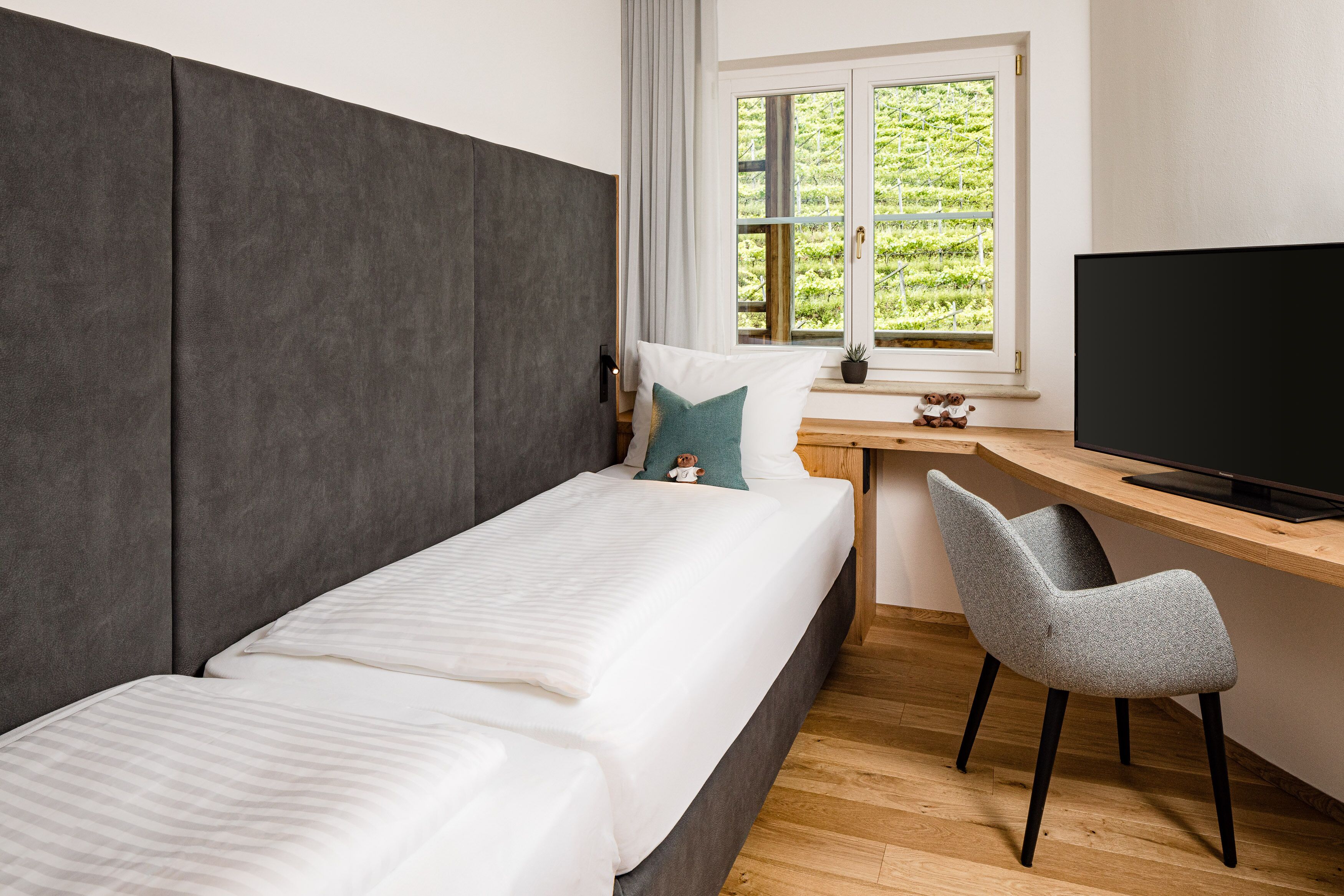A bedroom with a bed, a desk, a chair, a TV, and a window with a view of greenery.