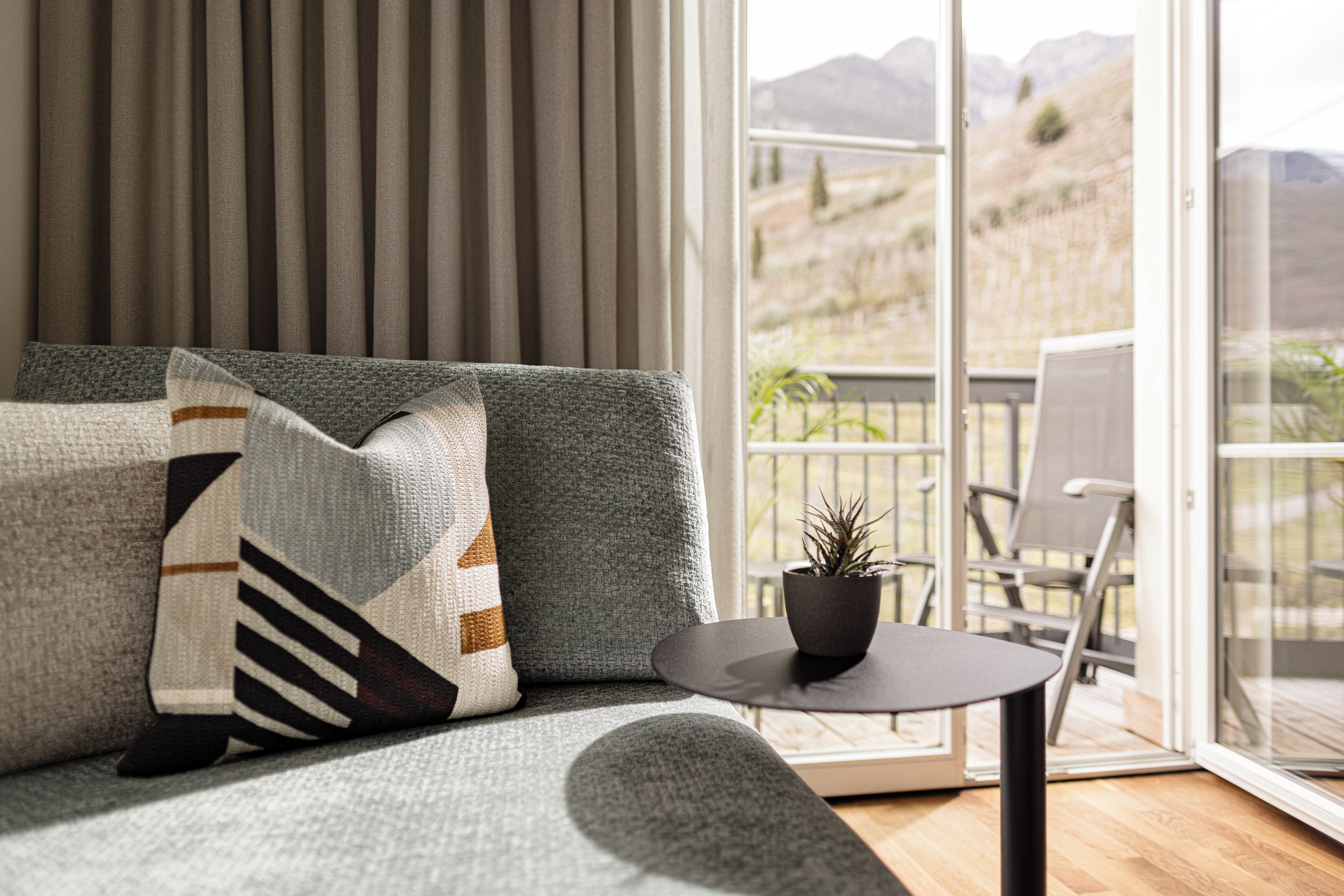 A modern living room with a grey sofa, patterned cushions, a small round table with a potted plant, and a sliding glass door leading to an outdoor balcony with a chair and a view of the countryside.