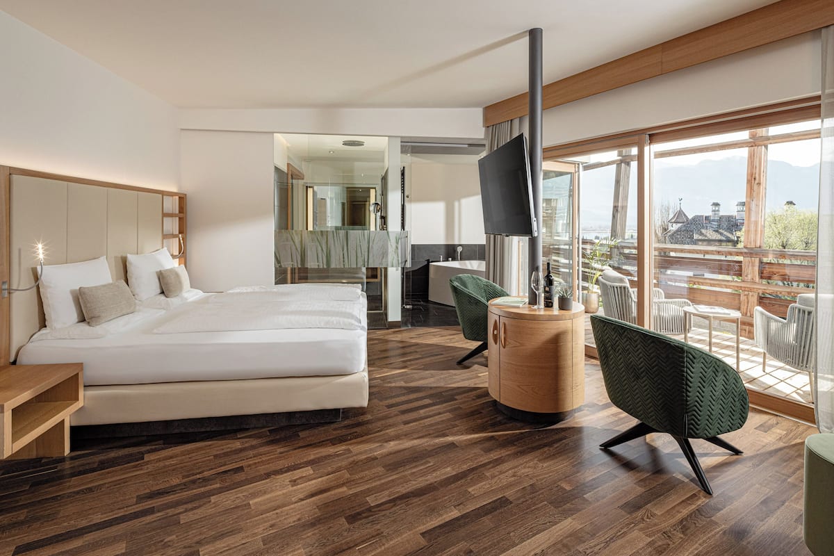 A modern hotel room with a large bed, a wooden floor, a green chair, a wooden side table, and a balcony with a view.