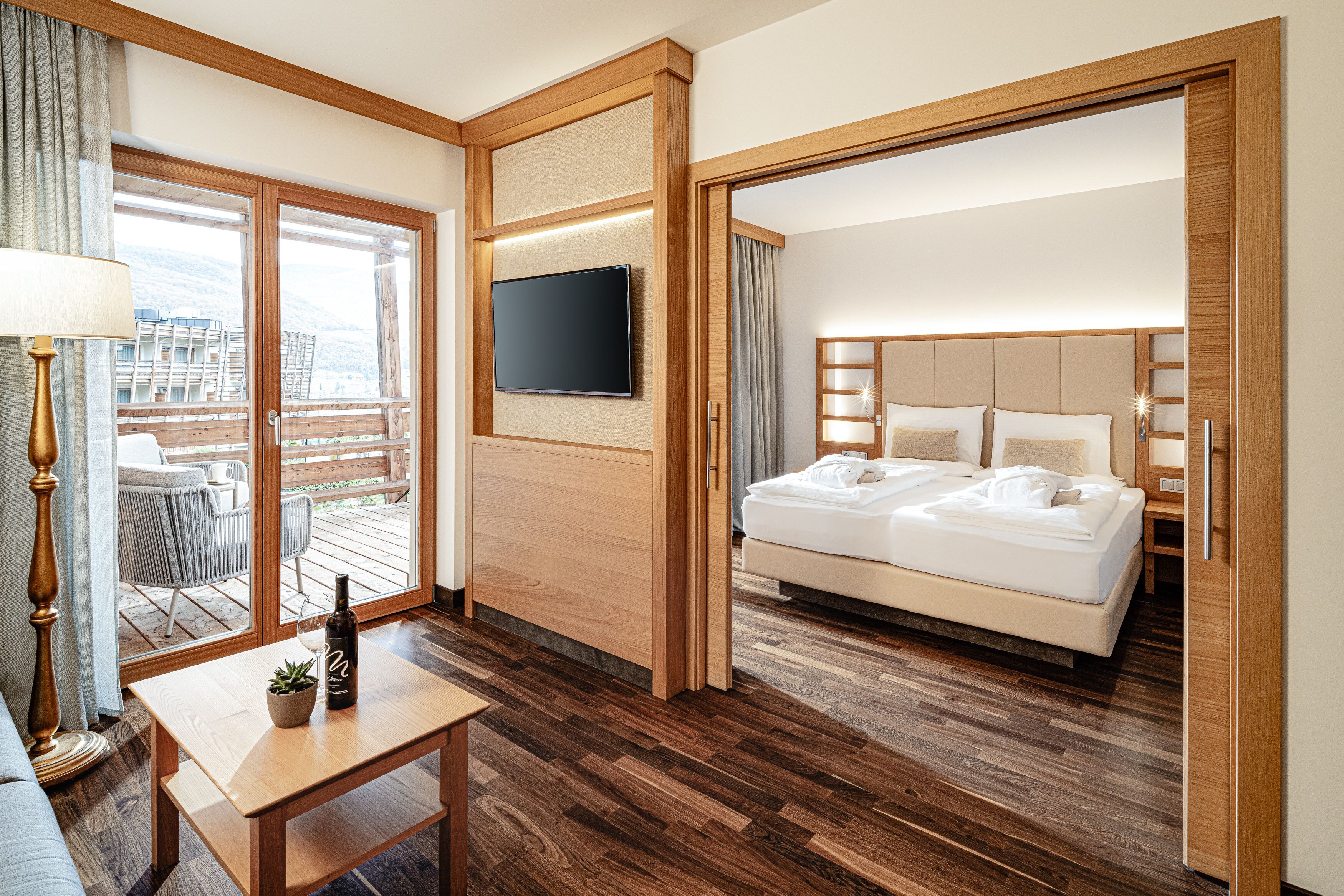 A hotel room with a bed, a TV, a wooden desk, and a balcony with outdoor furniture.