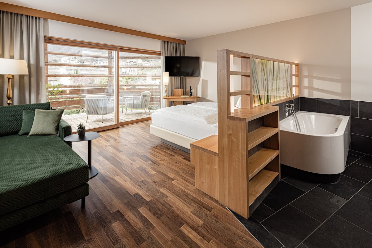 A modern hotel room with a bed, a sofa, a desk, a TV, a large window, and a bathroom with a bathtub.