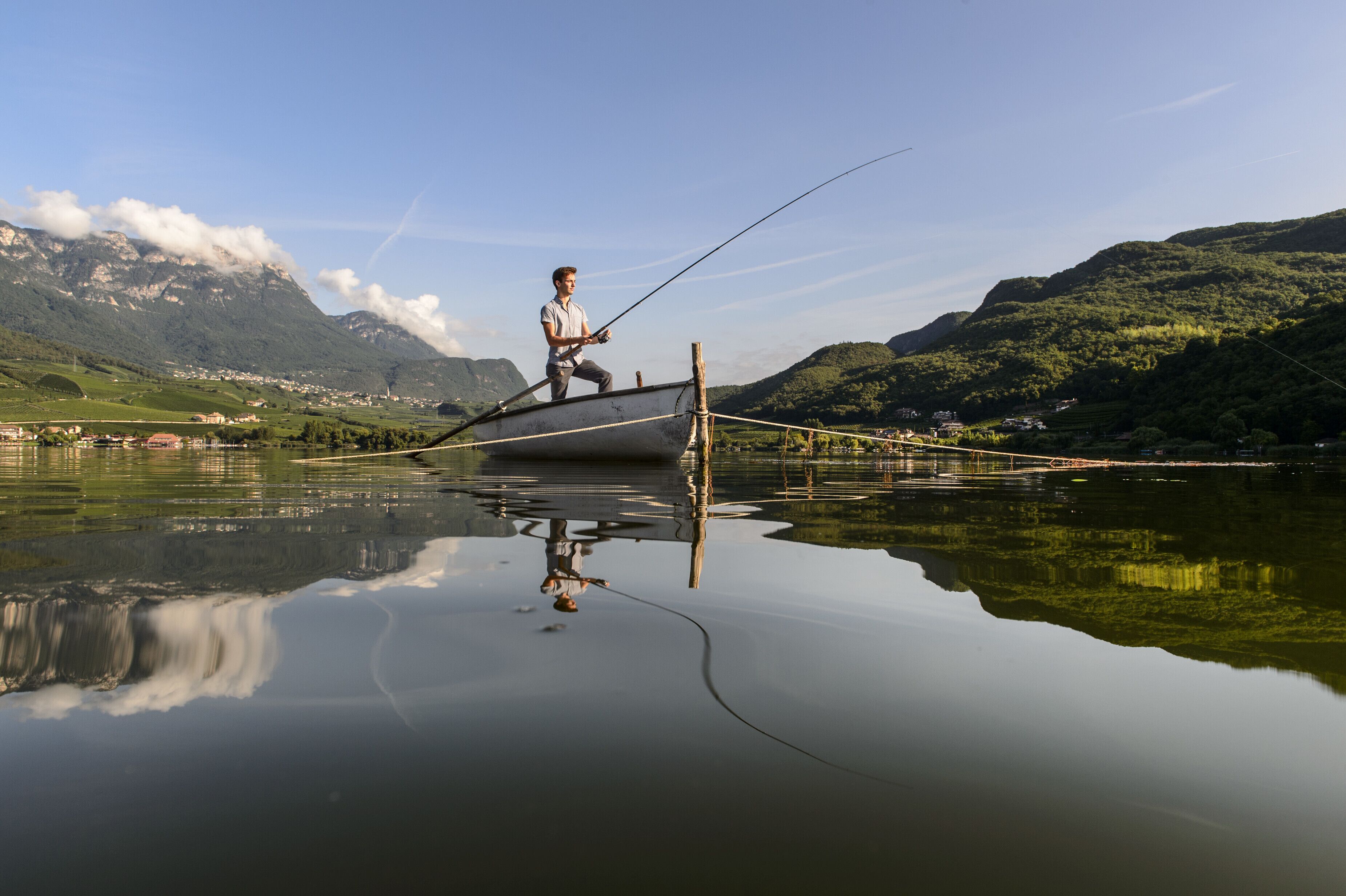 A person fishing on a boat in a calm lake with mountains in the background.