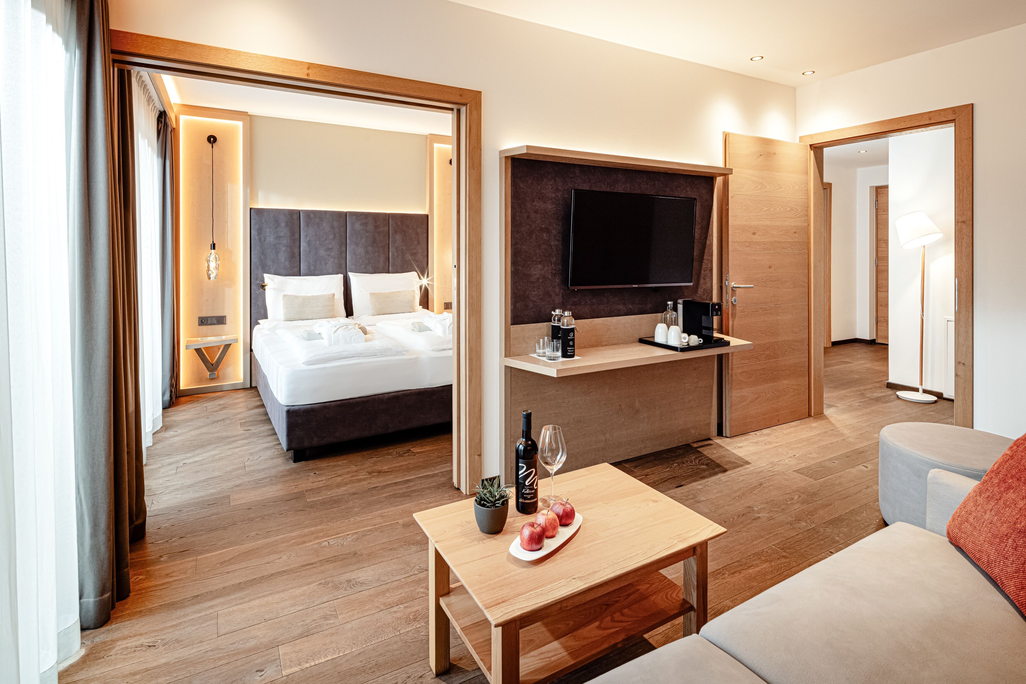 Ein modernes Hotelzimmer mit Bett, Wandfernseher, einem kleinen Tisch mit Flasche und Gläsern sowie einer Couch.