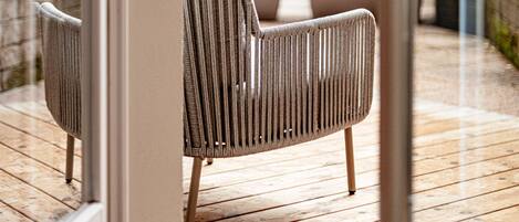 Een houten terras met een stoel en een tafel, een glazen deur die leidt naar een buitenruimte met groen.