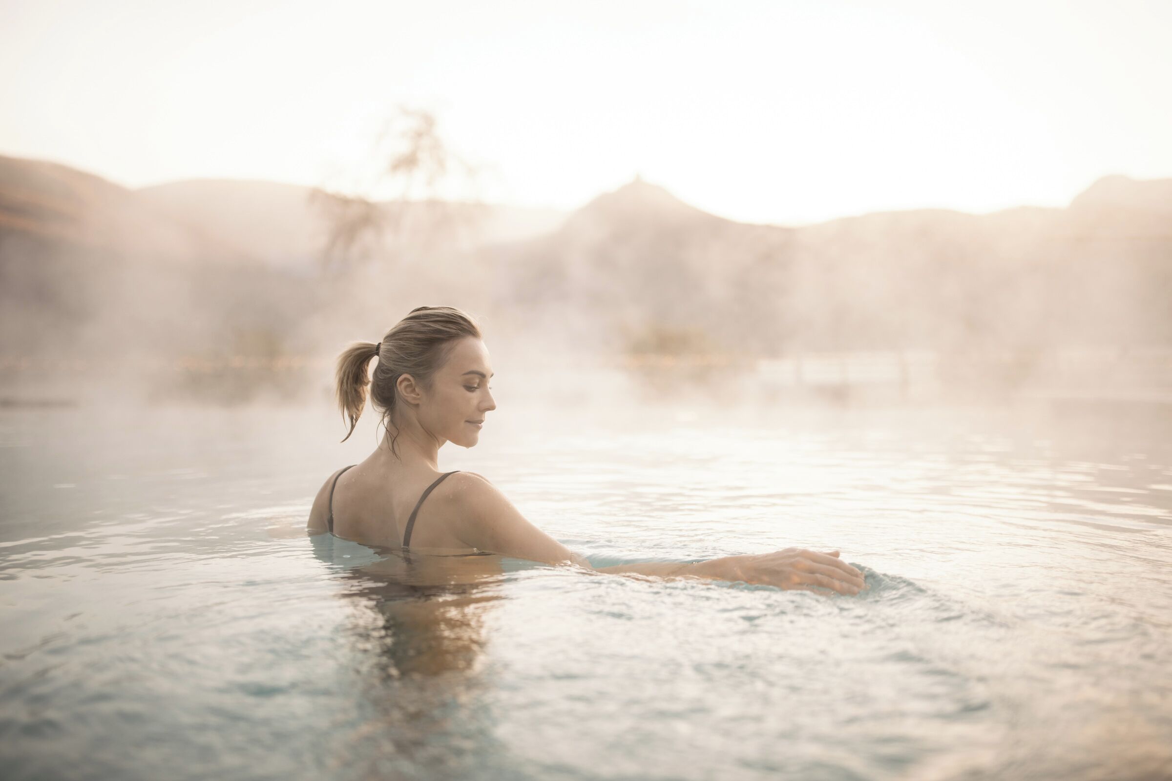 A person in a hot spring with steam rising from the water, surrounded by a mountainous landscape.