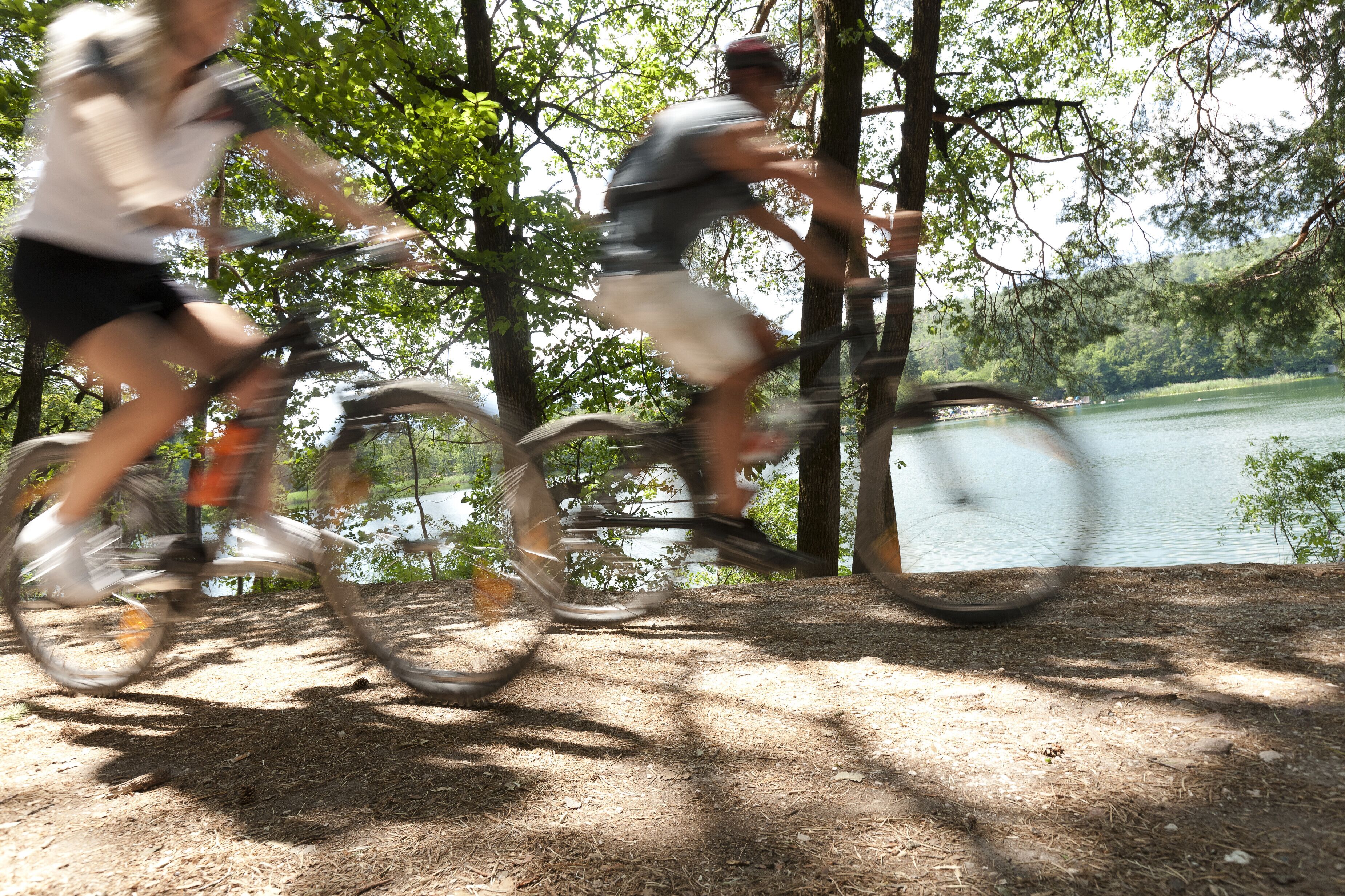 Two people riding bicycles on a dirt path with trees and a lake in the background.