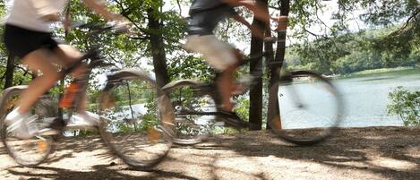 Two people riding bicycles on a dirt path with trees and a lake in the background.
