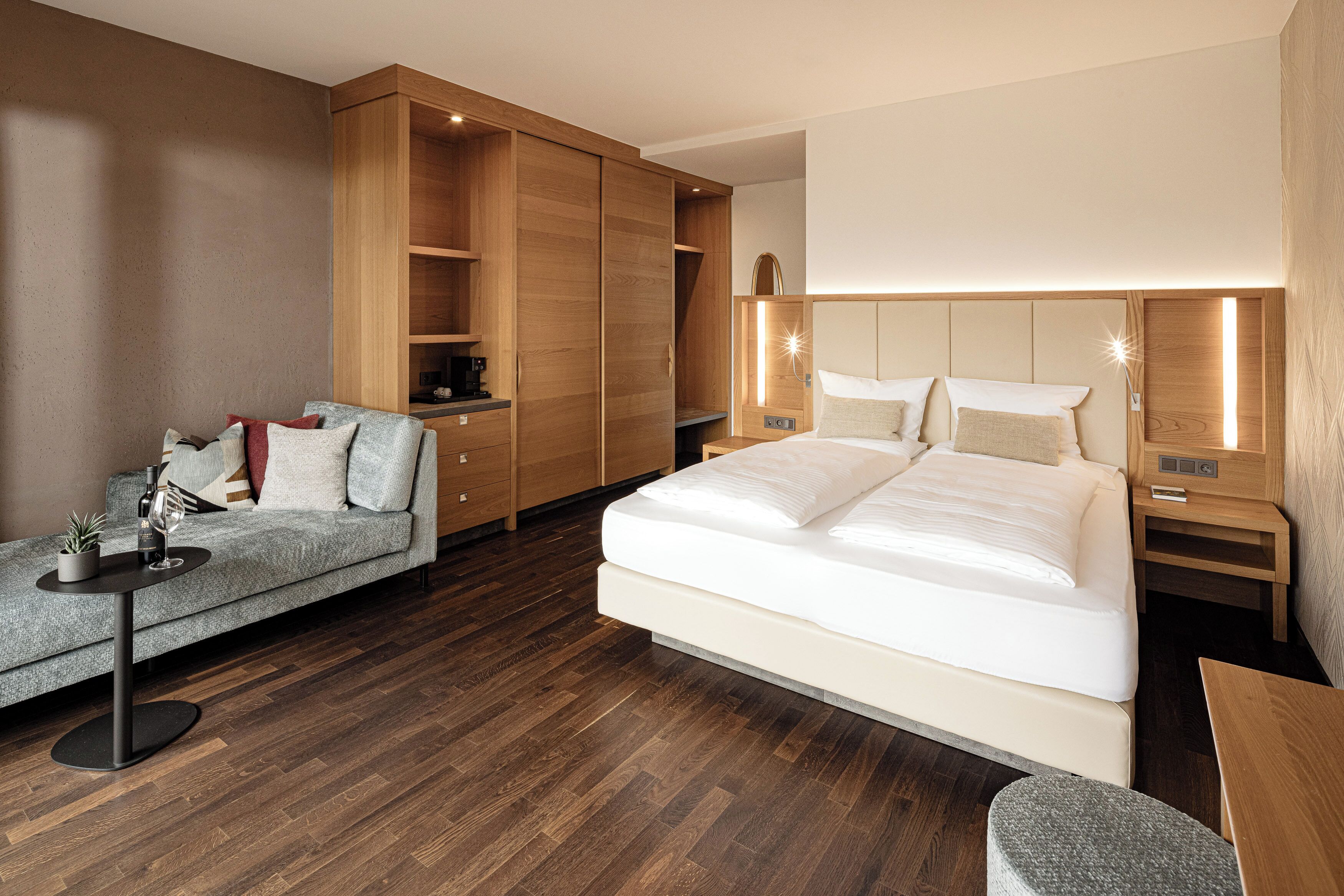 A modern hotel room with a large bed, a sofa, a wooden wardrobe, and a small table.