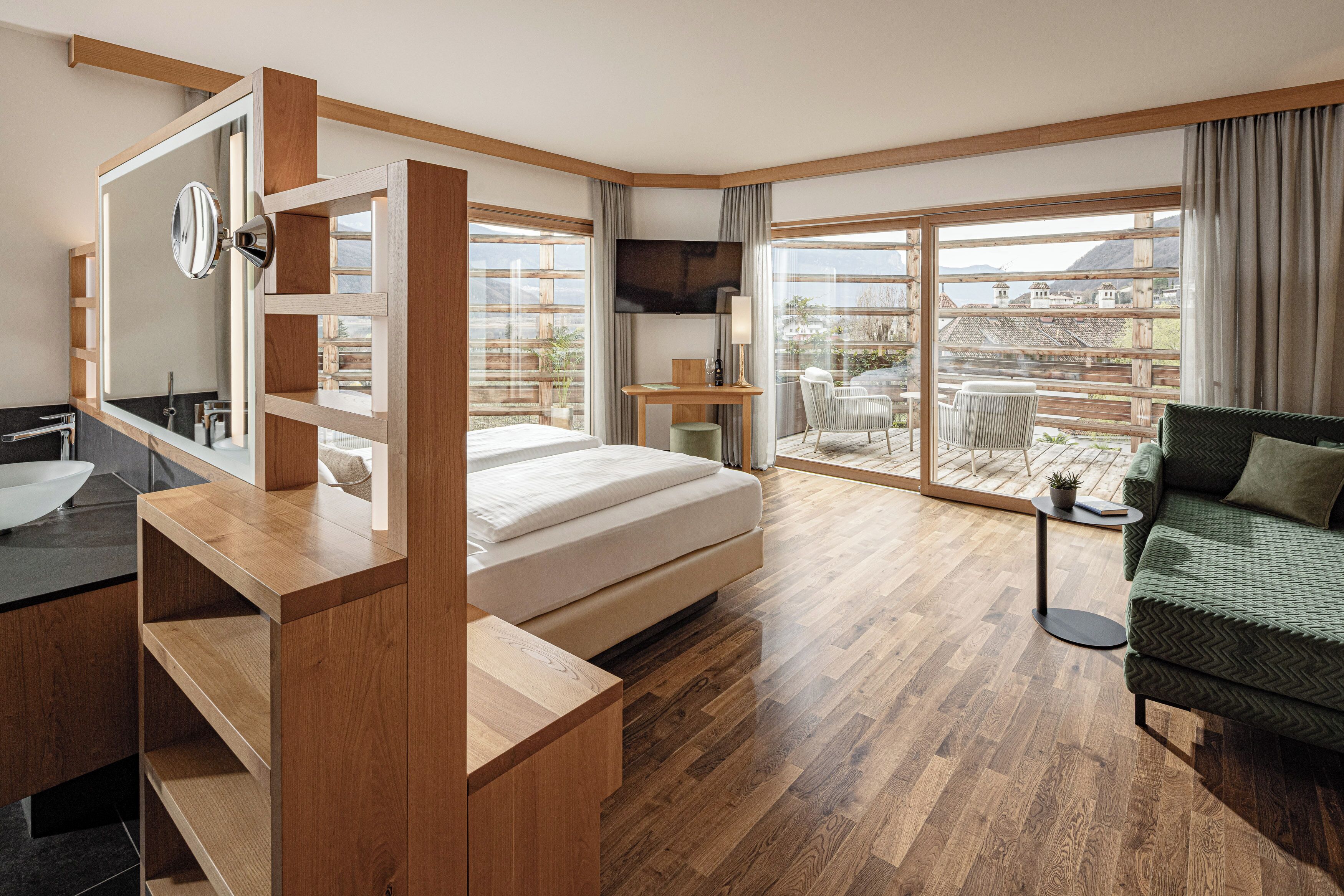 Una habitación de hotel moderna con piso de madera, una cama, un escritorio, un televisor, un balcón con vistas y un sofá verde.