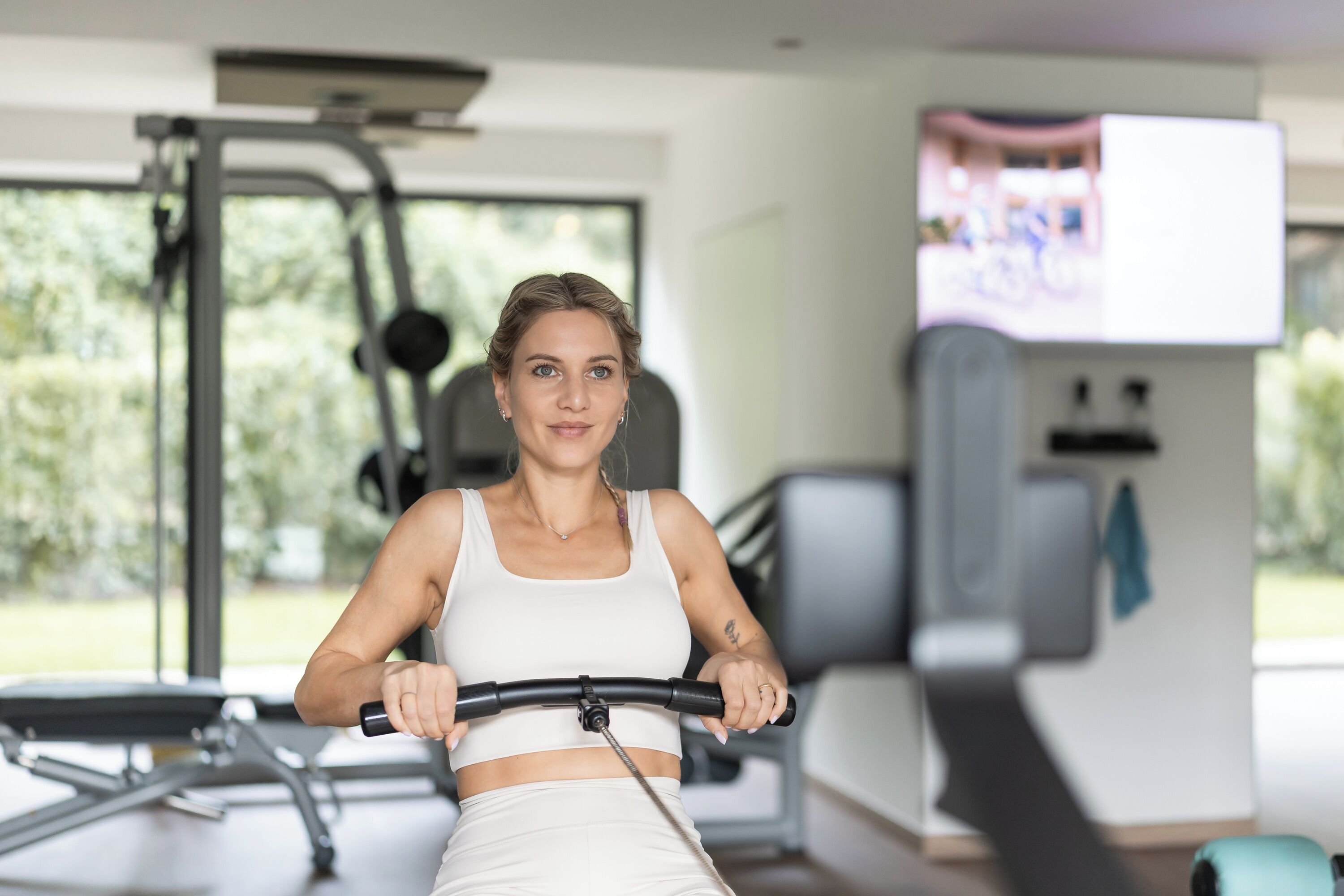 En kvinna tränar på en motionscykel i ett gym, med annan träningsutrustning och en tv-skärm i bakgrunden.