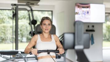 En kvinna tränar på en motionscykel i ett gym, med annan träningsutrustning och en tv-skärm i bakgrunden.