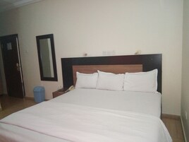 Habitación de hotel con una cama grande, cabecera de madera, mesita de noche con lámpara, espejo en la pared y un cesto de basura.