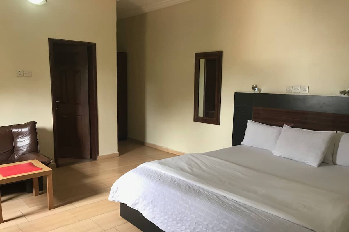 Un dormitorio con una cama grande, una mesita de noche de madera, un sillón de cuero marrón y una puerta que da a otra habitación.