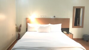 A neatly made bed with white linens, a wooden headboard, a bedside table with a lamp, and a small potted plant. - Bayview Resorts and Hotels (Enugu)