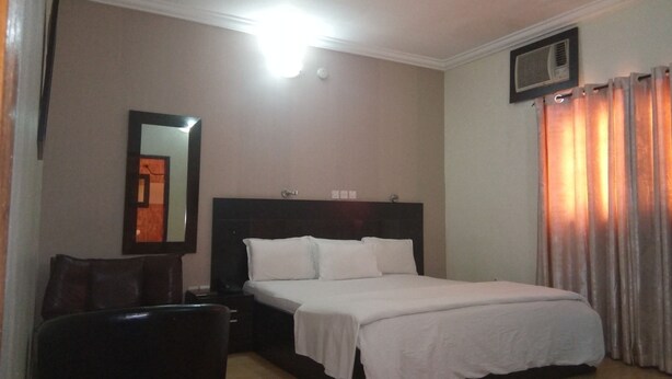 A hotel room with a bed, a nightstand, a chair, a mirror, and a window with curtains. - Bayview Resorts and Hotels (Enugu)