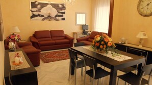 Ein Wohnzimmer mit einer Couch, einem Esstisch mit Stühlen, einer Wanduhr und einer Blumenvase.