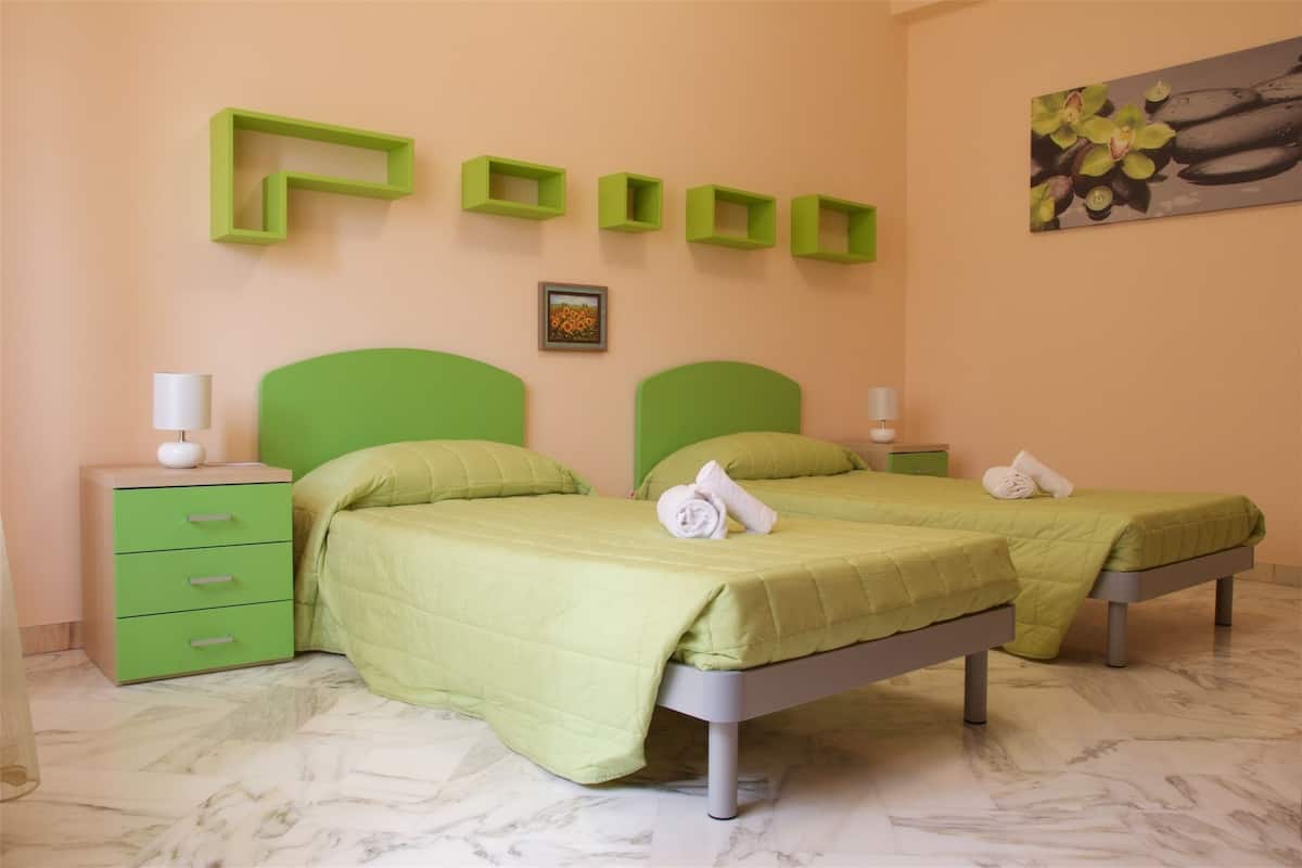 Een kamer met twee bedden, groene hoofdeinden, een nachtkastje in het groen, een witte lamp en een ingelijste foto aan de muur.
