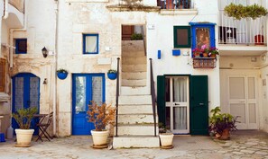 Une rue étroite bordée d’immeubles aux portes et fenêtres bleues, avec un escalier et des plantes en pot.