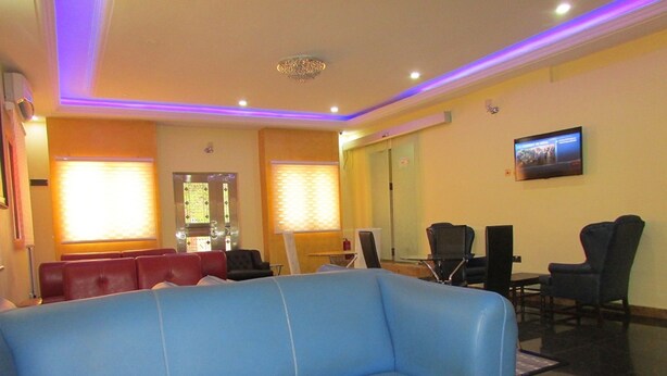 Un hall de hotel con un sofá azul, sillas rojas, una mesita y un televisor en la pared.