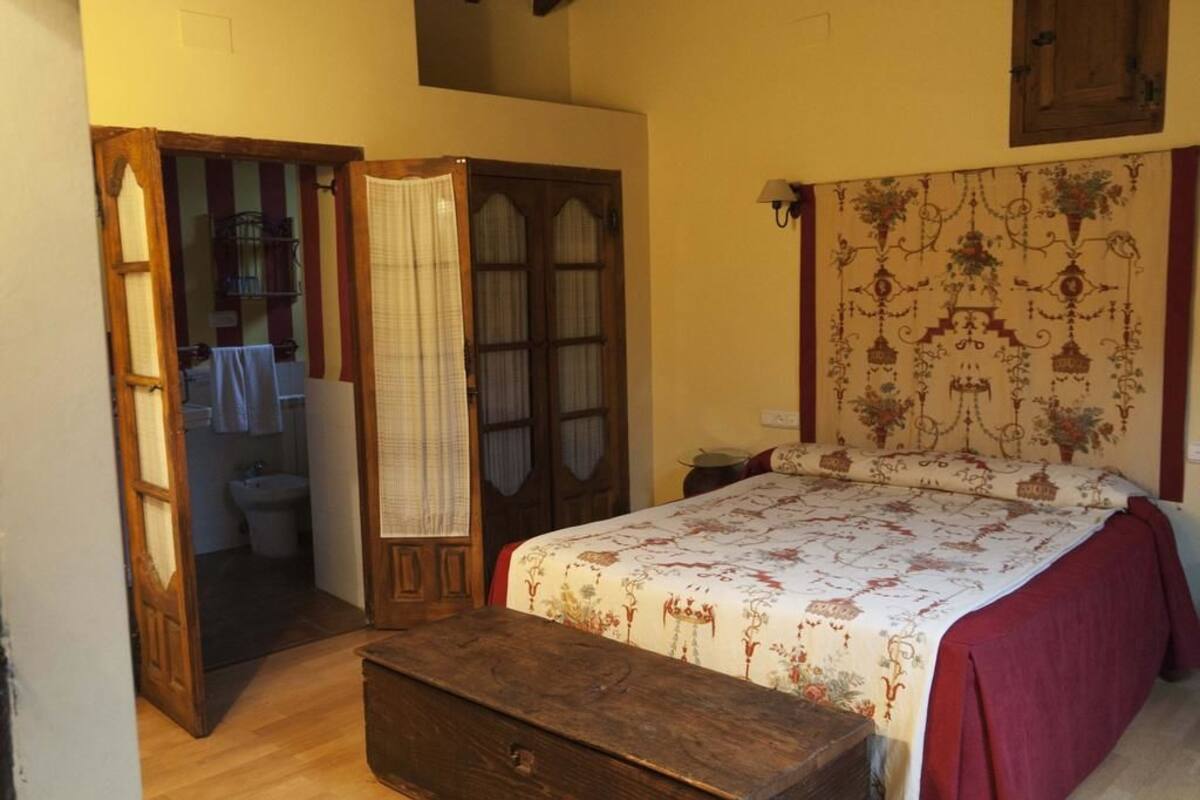 A bedroom with a wooden bed, a nightstand, a wooden chest, a bathroom with a sink and toilet visible through an open door, and a patterned headboard.