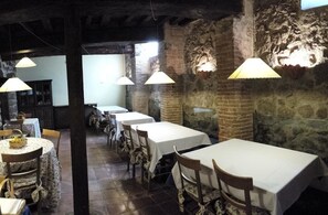 Un restaurant avec de longues tables recouvertes de nappes blanches, des lampes suspendues et des murs en pierre.