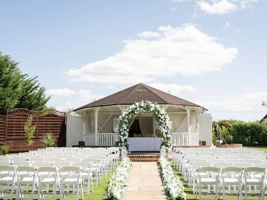 Montaje para una ceremonia de boda al aire libre con sillas blancas dispuestas en filas, un escenario techado y un arco floral.