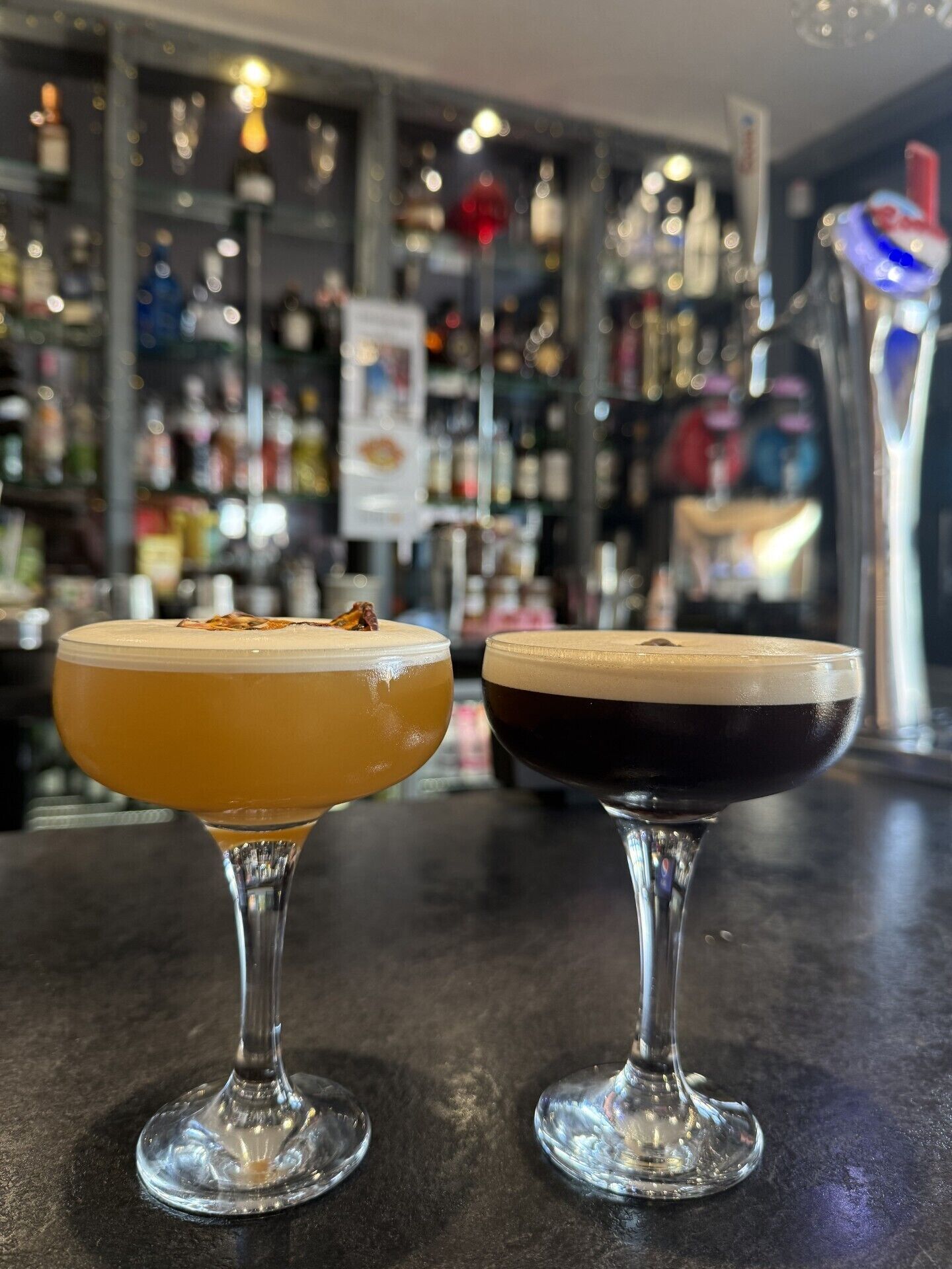 Dois cocktails sobre um balcão de bar, com um fundo desfocado de prateleiras e garrafas.