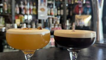 Dois cocktails sobre um balcão de bar, com um fundo desfocado de prateleiras e garrafas.