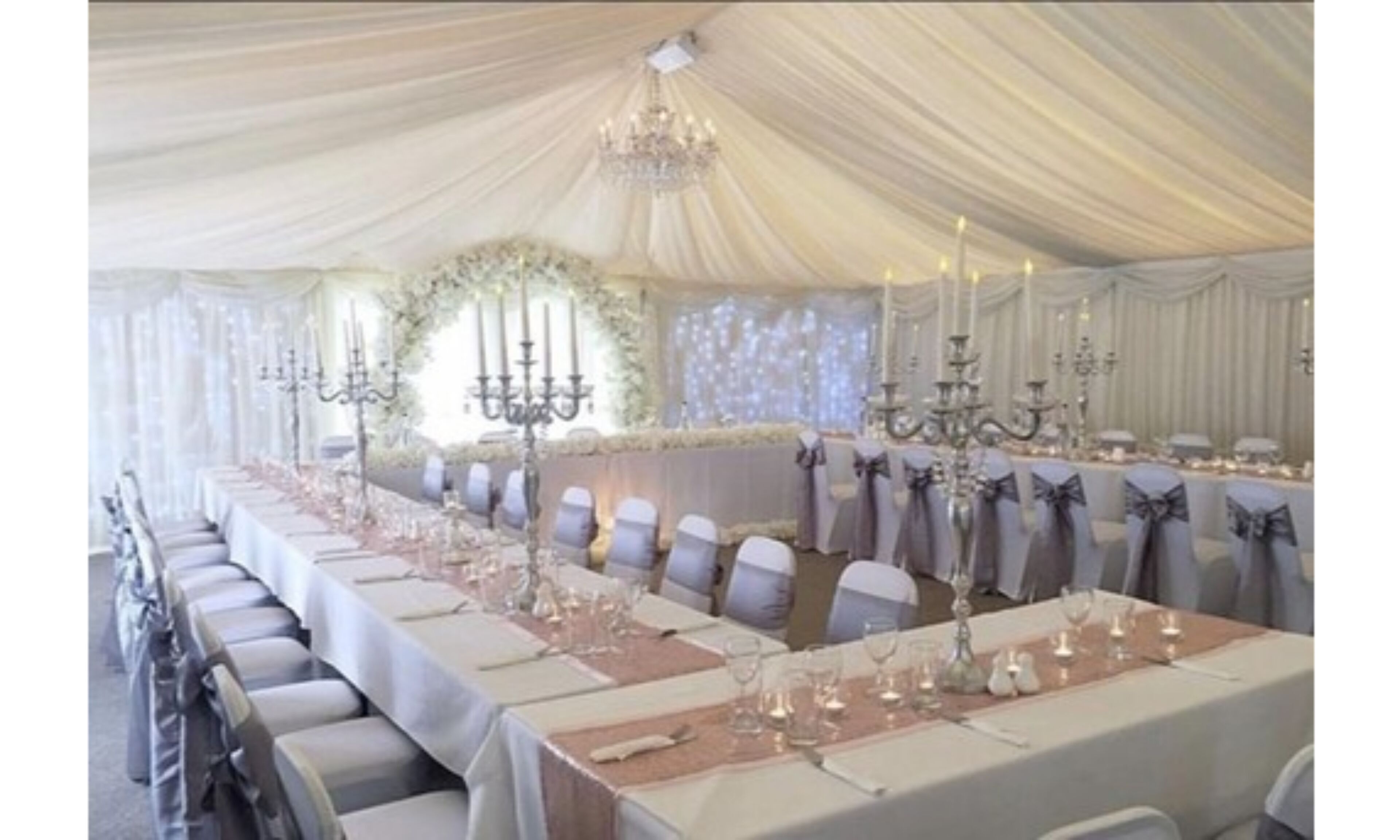 Un espace événementiel aménagé sous une tente, avec des tables dressées pour une occasion formelle, agrémenté de lustres et de compositions florales.