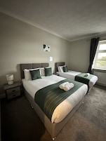 Quarto de hotel com duas camas, uma mesinha de cabeceira, uma luminária e uma janela com cortinas.