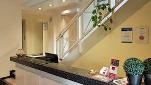 A hotel reception area with a counter, a clock, and a staircase with a plant. - Hotel Kronprinz (Minden)
