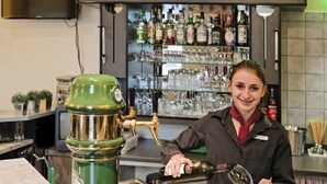 A bar area with a bartender preparing a drink, a green cocktail shaker, and shelves stocked with bottles and glasses. - Hotel Kronprinz (Minden)