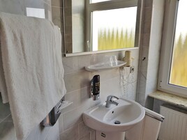 Un baño con un lavamanos blanco, un espejo, un perchero para toallas y un ventanal con vistas a árboles.