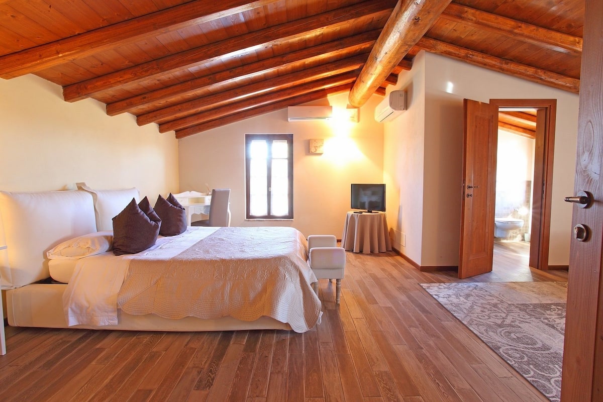 Een slaapkamer met een houten plafond, een bed met kussens, een bureau, een televisie en een badkamer zichtbaar door een open deur.