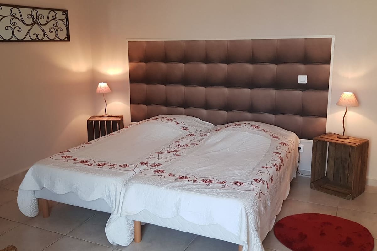 Une chambre à coucher avec une tête de lit capitonnée, un lit recouvert de linge de lit blanc orné de motifs floraux rouges, deux tables de chevet avec des lampes et un tapis rouge sur le sol.