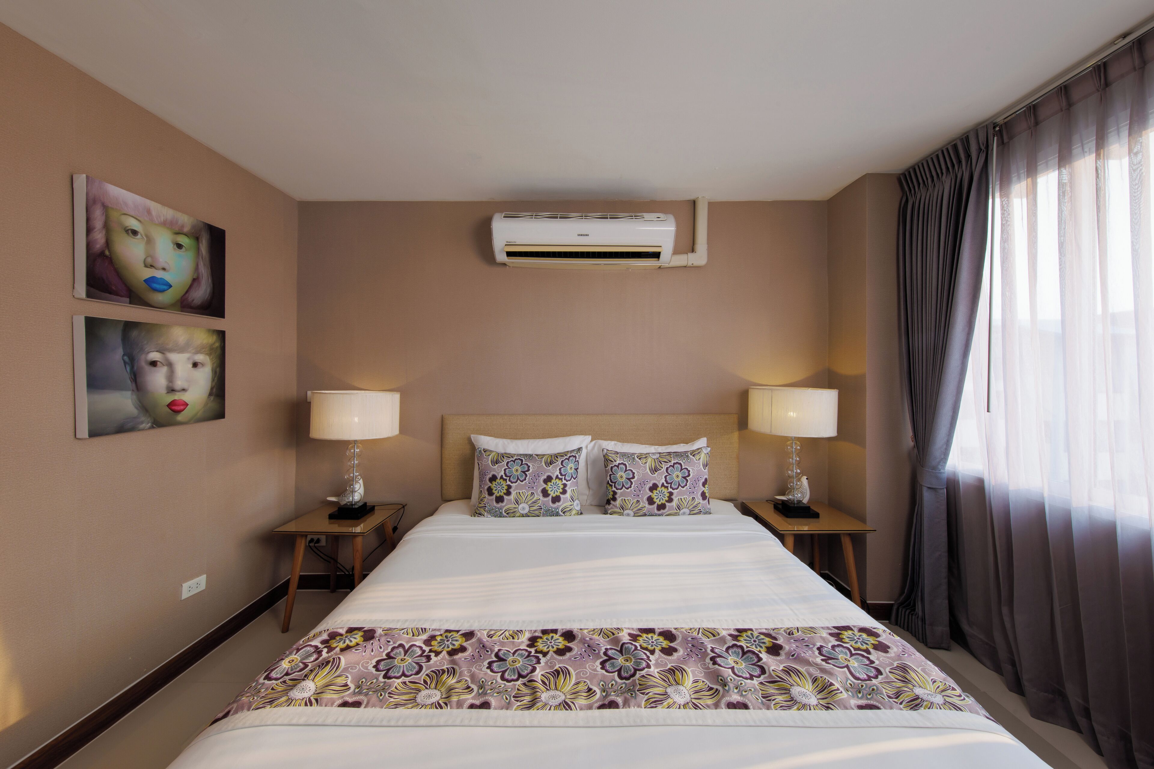 Une chambre d’hôtel avec un grand lit, deux tables de chevet avec des lampes, un appareil de climatisation et des œuvres d’art accrochées aux murs.