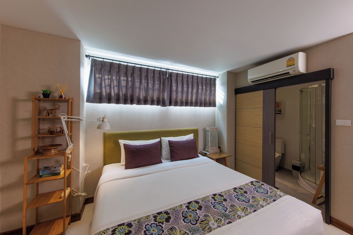 A hotel room with a bed, a bedside lamp, a shelf with decorative items, a window with curtains, and an air conditioning unit.