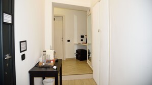 Un pasillo estrecho con suelo de madera, una mesa negra con un cesto de basura y una puerta que da a una habitación con un bolso negro y un pequeño estante.