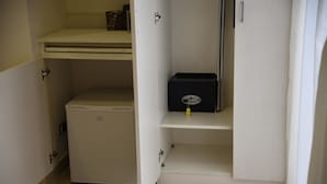 Una habitación pequeña con un mueble blanco compacto, un refrigerador blanco y una caja negra sobre una estantería.