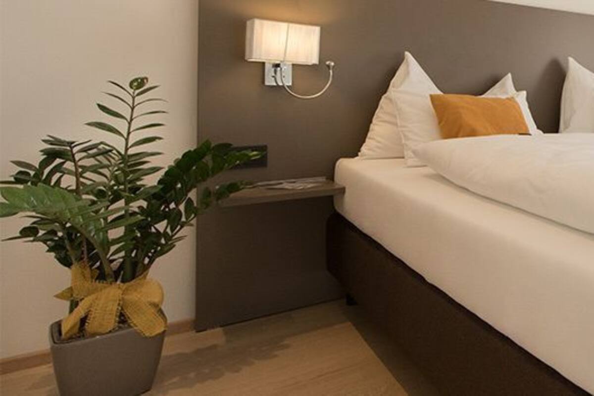 A hotel room with a large bed, two bedside tables, a nightstand, a potted plant, and two wall-mounted lamps.