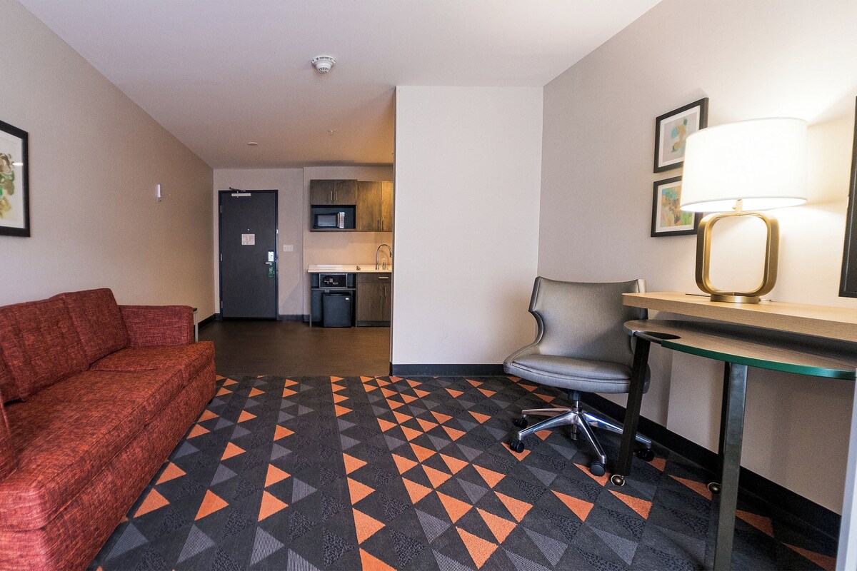 A hotel room with a red sofa, a desk with a lamp, a chair, and a kitchen area in the background.