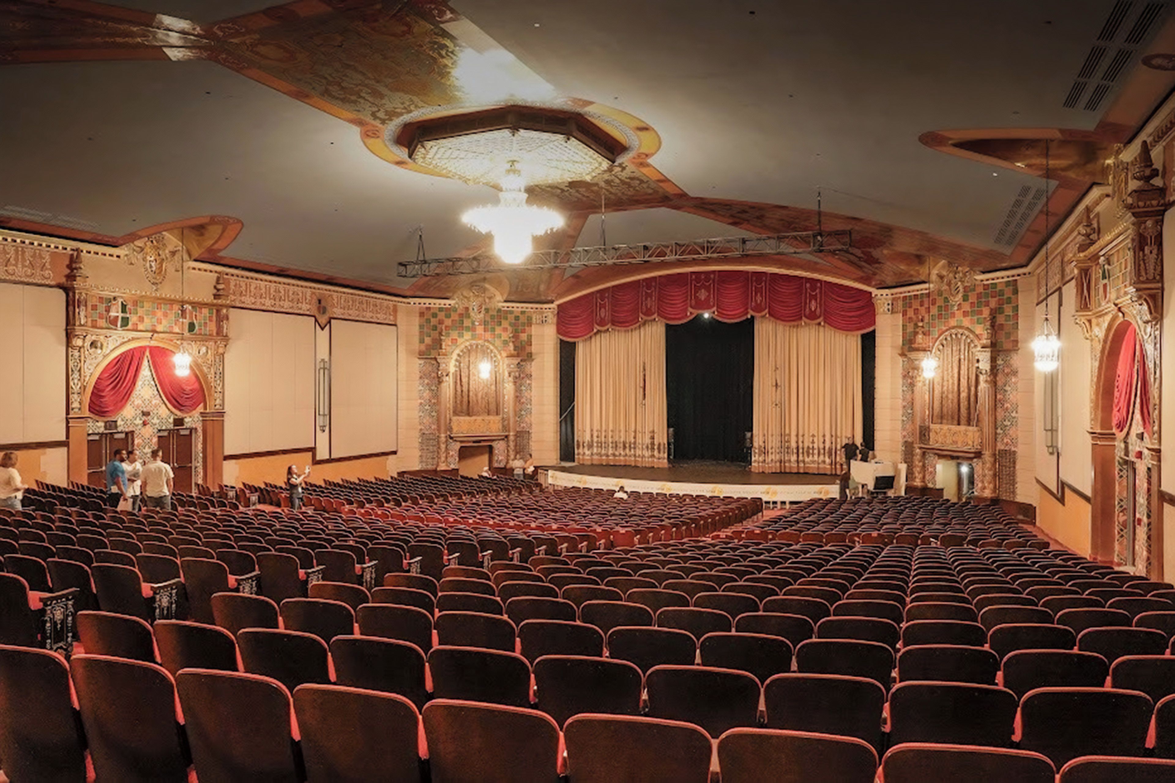 An auditorium with rows of seats, a stage with curtains, and ornate architectural details.