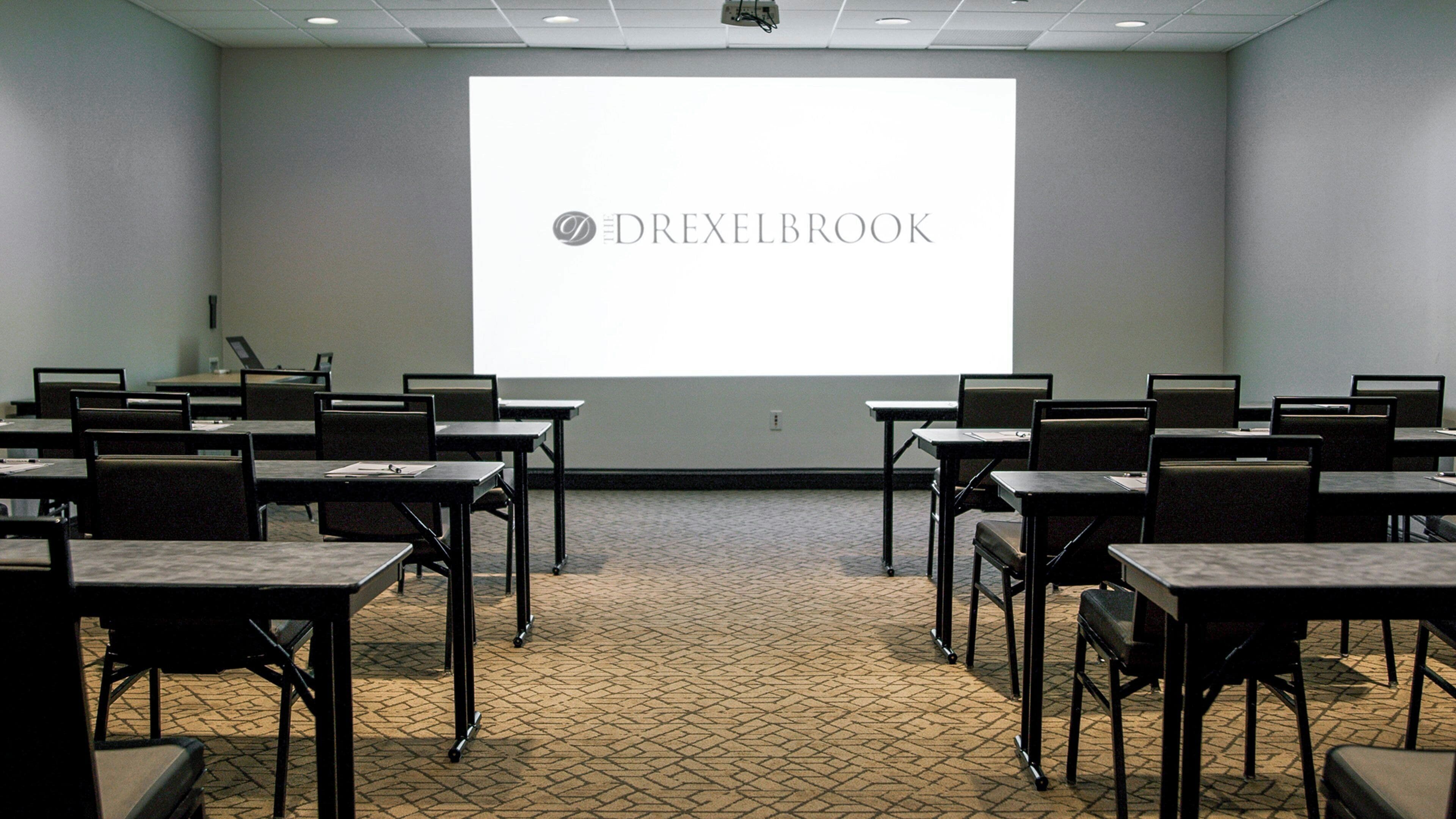 Un salón de conferencias con una pantalla de proyector mostrando 'DREXELBROOK' y varias mesas con sillas.