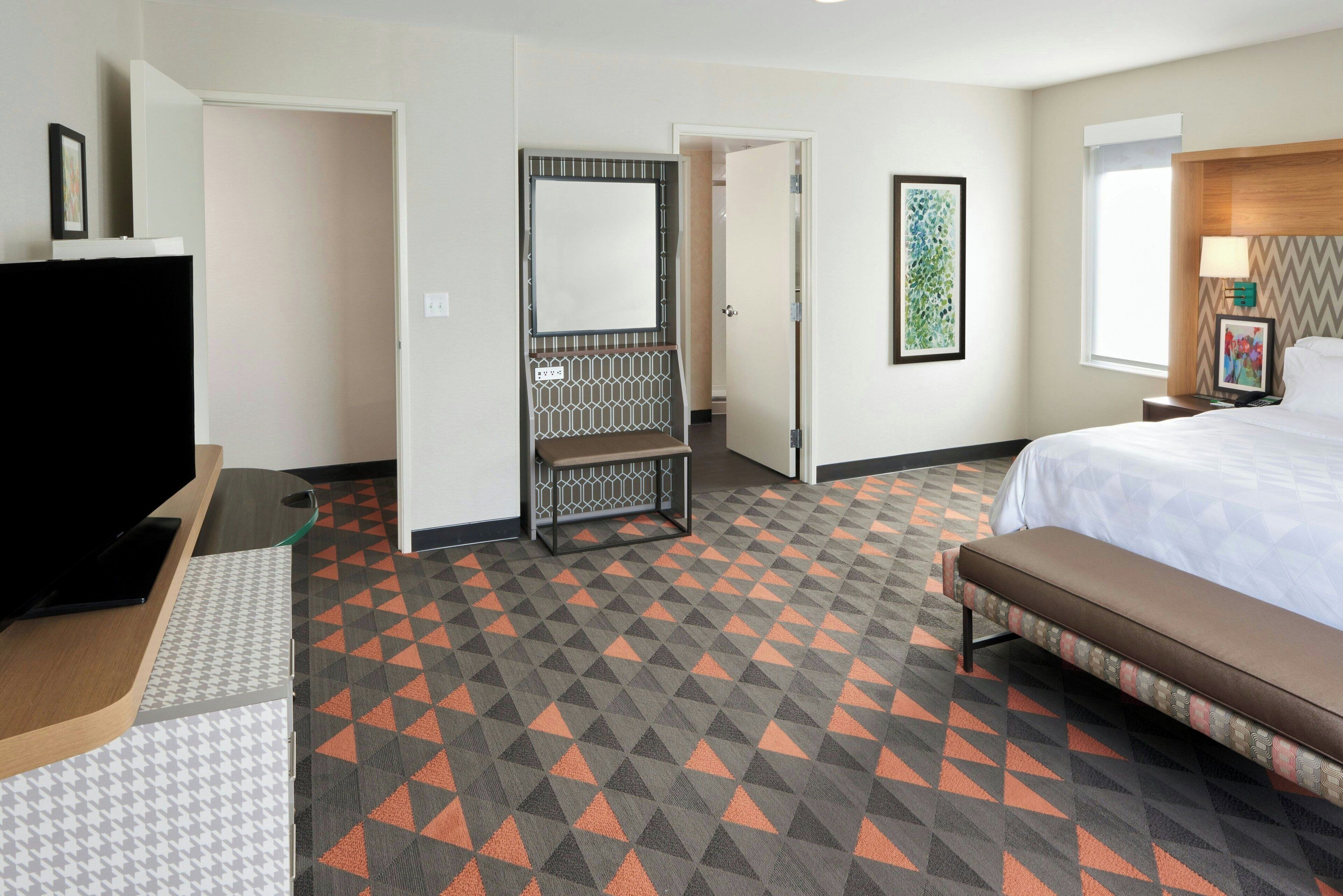 A hotel room with a bed, a desk with a flat-screen TV, a seating area, and a door leading to a bathroom.