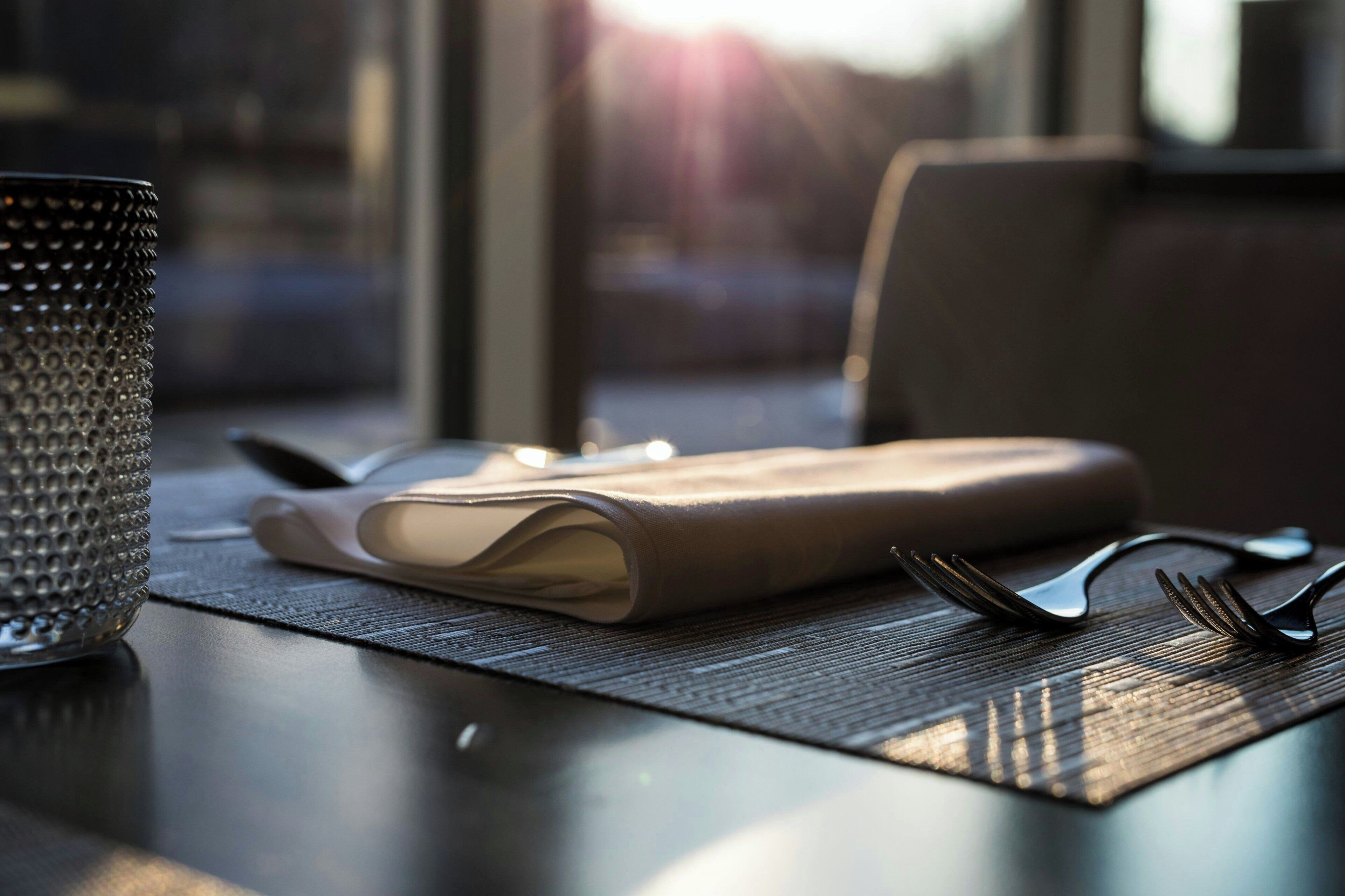 A table with a folded napkin, cutlery, and a textured container, likely a mug, set on a placemat.