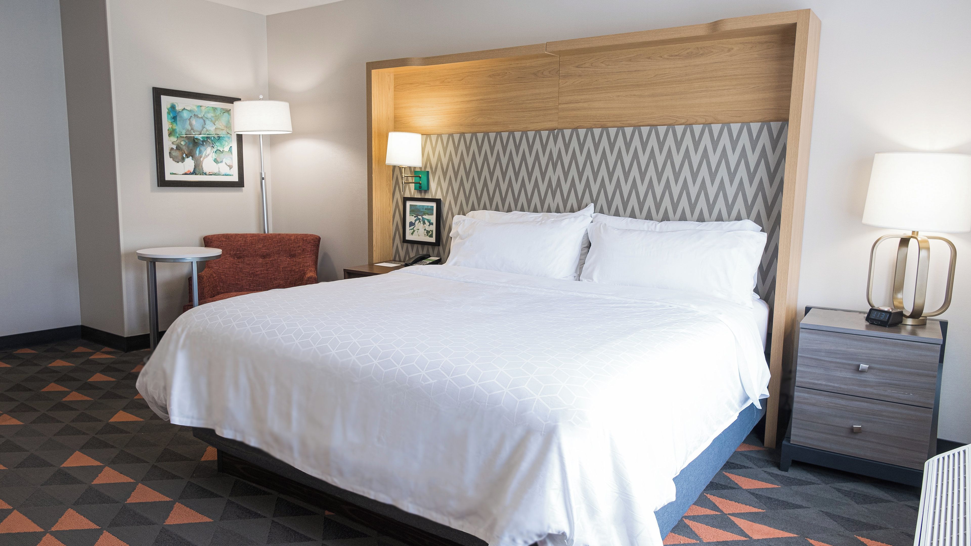 Quarto de hotel com uma cama grande, mesa de cabeceira, luminária de parede e uma cadeira.