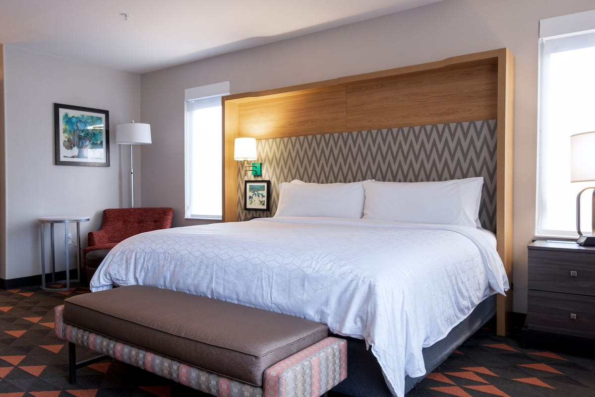 A hotel room with a large bed, a wooden headboard, a bench, a chair, and a desk with a lamp.