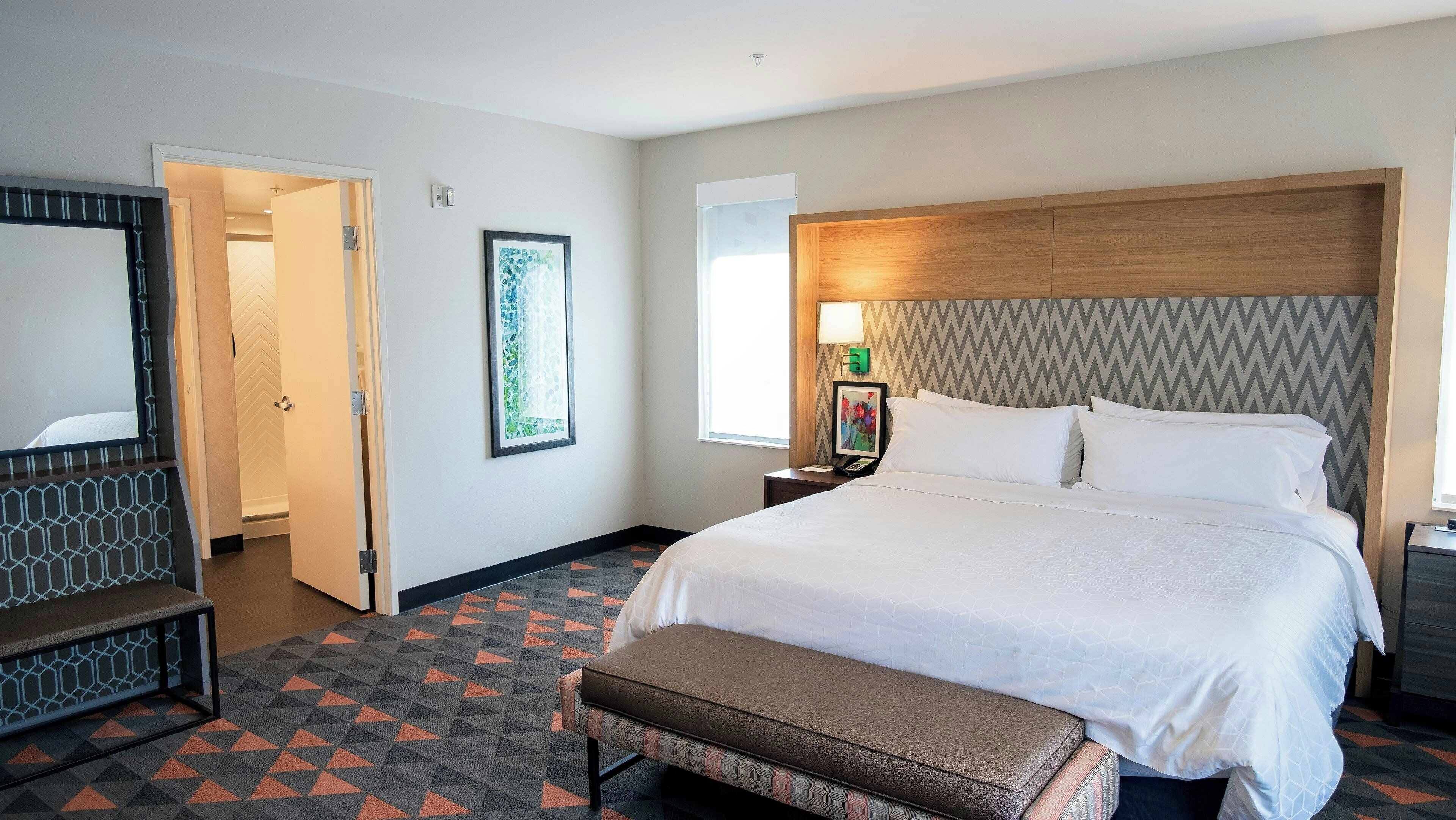A hotel room with a large bed, a bench, a desk with a lamp, and a picture on the wall.