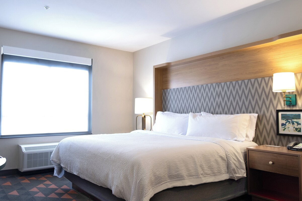 A hotel room with a bed, bedside lamps, a nightstand, a desk, and a window with blinds.
